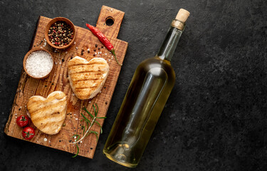 
two heart shaped grilled chicken breast steaks with spices, a bottle of white wine for Valentine's Day dinner on a stone background with copy space