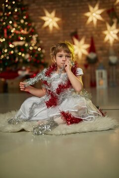Children portrait for Christmas - little girl in white dress in front of Christmas tree at home. Christmas lights in the background.