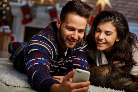 Christmas couple at home in Winter. Happy young couple lying on floor using smart phone at home with dog at Christmas time. Christmas tree and fireplace in background.
