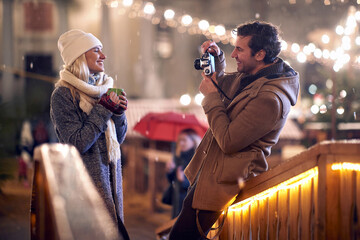 young man taking a photo of his girlfriend holding cup of tea outdoor with retro camera