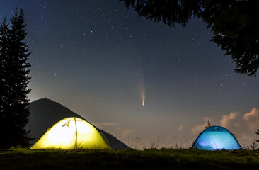 Fototapeta na wymiar Two brightly lit tourist tents on forest clearing in mountains with starry sky and C/2020 F3 (NEOWISE) comet with light tail.