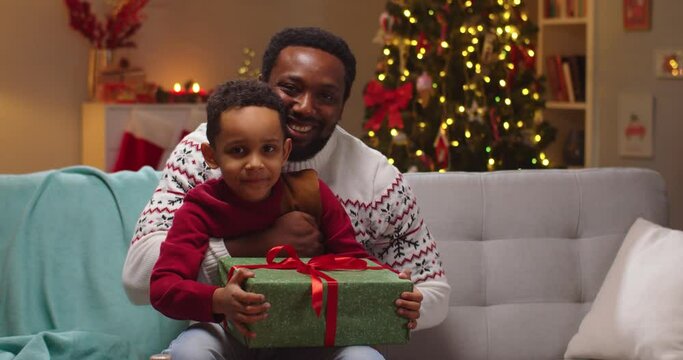 Close up portrait of African American happy caring dad hugging, embracing little cute son with xmas gift sitting in room with glowing decorated tree and spend time together on Christmas Family concept