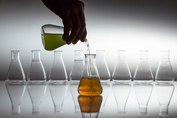 Hand pouring green in to orange liquid in scientific laboratory glass erlenmeyer flask with glassware equipment on reflective surface.
