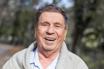 Fototapeta premium Portrait of happy senior man smiling, in the public park, outdoors. Old man relaxing outdoors and looking at camera. Portrait of elderly man enjoying retirement