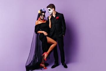 Dark-haired beautiful bride and groom with carnival makeup posing in black dresses on isolated background