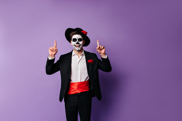 Positive dark-haired guy in Mexican-style clothes joyfully shows his fingers upwards, posing for portrait with space for text