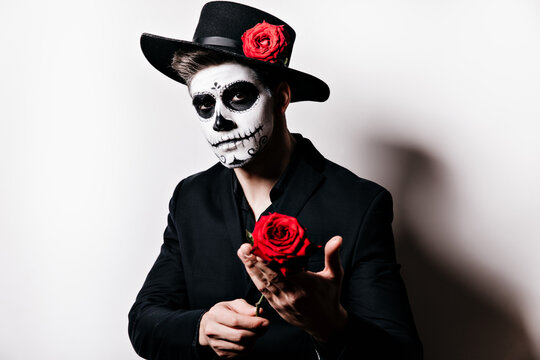 Close-up shot of romantic guy with painted face in form of skull, presenting red rose