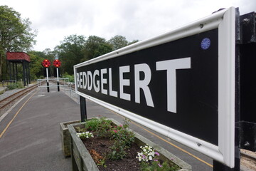 A sign of Beddgelert station in Wales from the side, with the platform end in the background in grey weather.