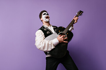 Face art guy imagines himself rock musician and emotionally poses with guitar. Indoor portrait of man on lilac background