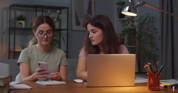 Portrait of Caucasian twin sisters sitting in room and speaking while using devices. Female typing on laptop while her twin searching internet on smartphone and showing her something. Leisure concept