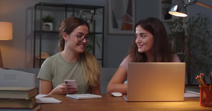 Portrait of cheerful Caucasian twin sisters sitting at home and speaking while using devices. Beautiful female typing on laptop while her twin browsing on smartphone and chatting. Leisure concept