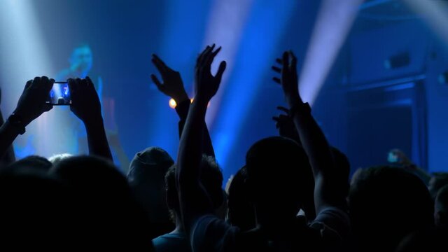 Slow motion: people crowd partying, cheering, raising hands up and clapping at rock concert in front of stage of nightclub. Bright colorful stage lighting. Nightlife and entertainment concept