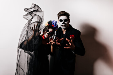 Portrait of young guy and girl in black clothes and frightening masks posing like zombies against background of their own shadows