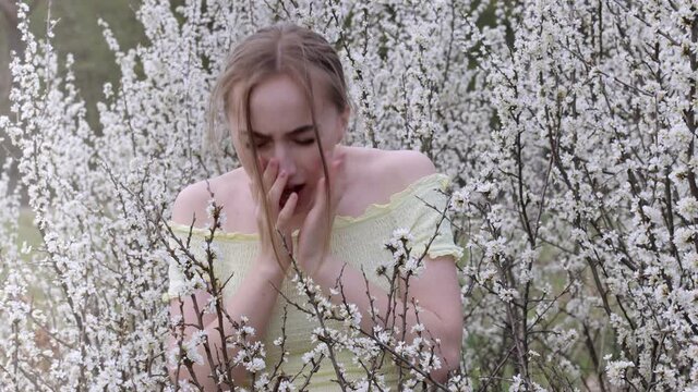 Young woman blowing nose and sneezing in tissue in front of blooming tree. Seasonal allergens affecting people. Beautiful lady has rhinitis