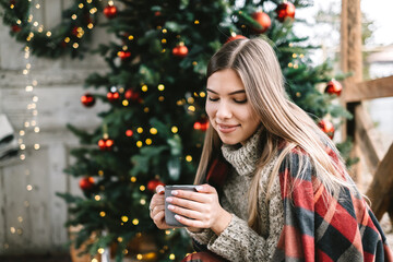 Young caucasian woman in a plaid is drinking a hot drink on the veranda with Christmas tree and decorations.