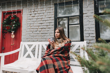 Young caucasian woman in a plaid is drinking a hot drink sitting on a bench in the backyard.