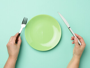 two hands hold a metal knife and fork on the surface of a round empty green plate