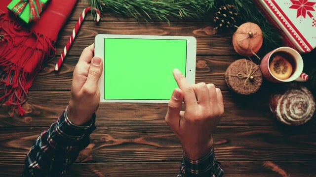 Man looking at photos on tablet holding device over wooden table with winter decor. Top view. Green screen.