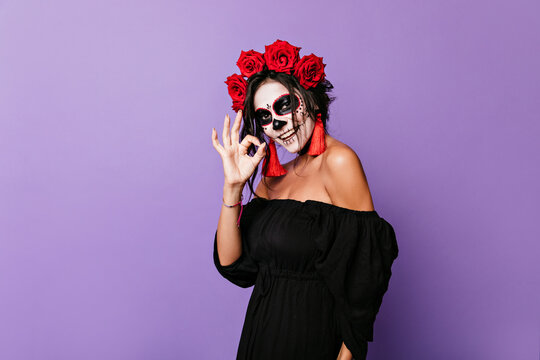 Charming girl in image of skeleton happily posing in studio. Portrait of cute lady in black top with red roses in curls showing OK