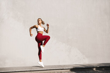 Fitness woman doing cardio interval training outdoors. Caucasian female in sportswear exercising outdoors in morning, jumping against concrete wall