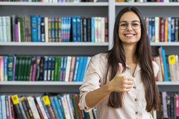 Beautiful female student looking at the camera and showing thumbs up against the background of the library bookshelves. Learning and education concept