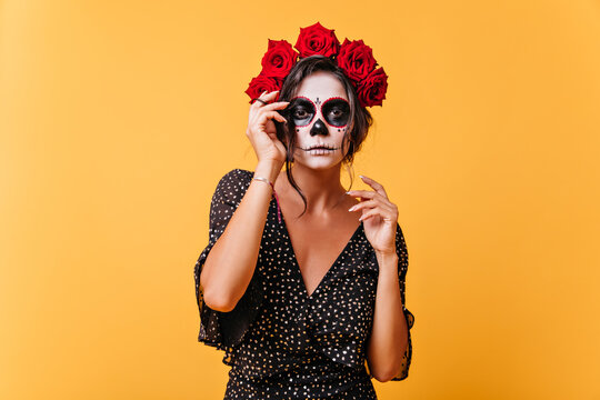 Dark-skinned girl with crown of flowers and mask of skull poses for photo on memory of Halloween. Portrait of extraordinary model in unusual outfit