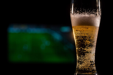 Close-up of fresh foaming light beer in a glass and football on TV on background. Sports bar concept