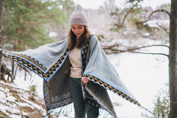 Young brunette beautiful woman in hat and grey poncho walking in the winter forest - obrazy, fototapety, plakaty