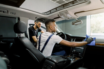 Car service and detailing station. Back angle indoor shot of two professional multiethnic men workers, providing a vehicle interior cleaning, wiping front panel with microfiber cloth and brush