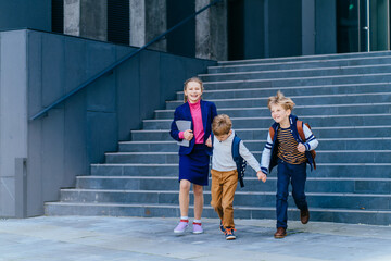 Obraz premium Happy pupil preteen girl and her two friends are going to school. Three different age kids running downstairs outdoors with school building on background. Happy childhood, back to school concept.