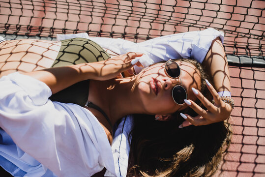 Stylish cute teenage model dressed in summer hipster vintage white stylish clothes. Trendy girl posing in sunglasses lying on a tennis court. The original shadow from the grid at the sports field