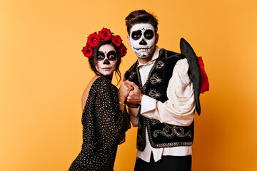 Portrait of pair of lovers on isolated orange background. Girl and guy in form of skeletons hold...