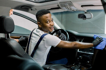 Professional car detailing service. Back angle view of cheerful African man worker, wiping front plastic panel with microfiber cloth, smiling to camera. His colleague cleans car door on the background