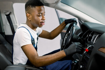 Auto service and professional detailing concept. Young black man worker in white t-shirt and overalls, makes vehicle interior cleaning, wiping front plastic panel with a brush