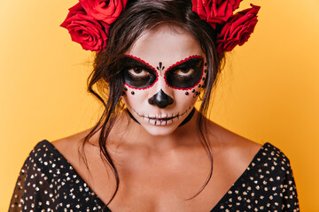 Close-up photo of brown-eyed woman with carnival face art. Mexican model is angry looking at camera on orange background