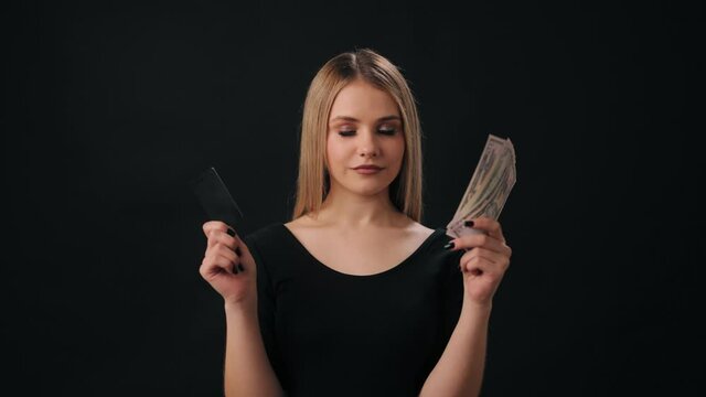 Portrait of young woman comparing cash and credit card that holding in hands. Happy blonde choosing better way of payment. Isolated over black background.