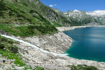 A torrent rushing towards a lake. The artificial lake stretches over a vast territory, shining with navy blue color. The dam is surrounded by mountains.A glacier in the back. Controlling the nature