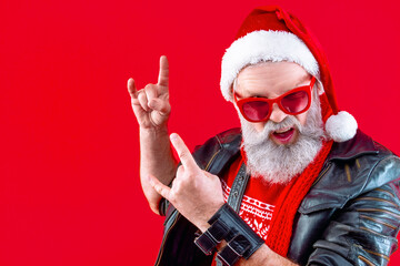 Fototapeta na wymiar Happy, old man with mustache and long white beard show rock hands. Modern Santa Claus wearing stylish red sweater, sunglasses, hat and lather jacket. Winter holidays, new year celebration concept