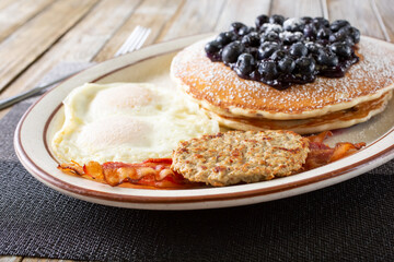 A view of a pancake breakfast platter, featuring over easy eggs, bacon strips and a sausage patty.
