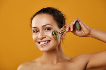Beautiful african woman smiling and massaging face skin with jade roller isolated on yellow