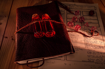 Image of a leather bound tome with on top two bottles with red liquid. Role playing dice, pencils...