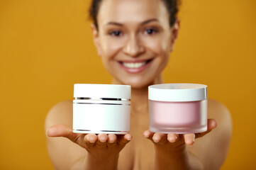 Fototapeta premium Beautiful african woman demonstrating jars of creams at camera isolated on yellow