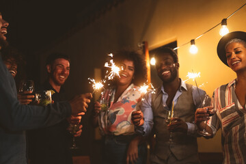 Happy young friends celebrating new year eve with sparklers fireworks and drinking cocktails on patio house party - Youth people lifestyle and holidays concept