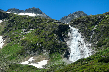 Fototapeta na wymiar A cascading waterfall in the Austrian Alps. The slopes around it are overgrown with lush green grass. The waterfall flows along very steep slope. Sunny and bright day. Power of the nature. Remedy