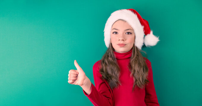 Happy Christmas girl in a red suit and a Santa Claus hat on a green background. Red and green. Portrait of child. The girl shows likes. Space for copying.