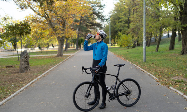 A young man in a bicycle helmet drinks water from a fitness bottle and holds a bicycle in his hands. Sports activities and food delivery around the city on bicycles.