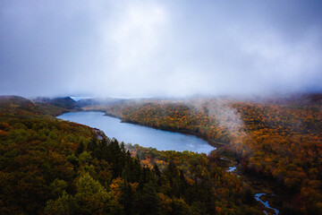 Beautiful travel aerial photograph of Lake of the Clouds Overlook as fog lifts from the trees below on an overcast morning with colorful autumn foliage blanketing the hills and valley below.
