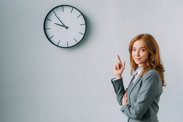  redhead businesswoman pointing with finger at wall clock, while looking at camera on grey