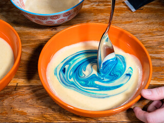 cooking sweet sponge cake at home - top view of stirring blue food coloring in bowl of liquid dough on old wooden table at home kitchen