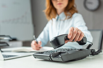 Cropped view of businesswoman putting handset on landline telephone, while sitting at workplace on blurred  - obrazy, fototapety, plakaty